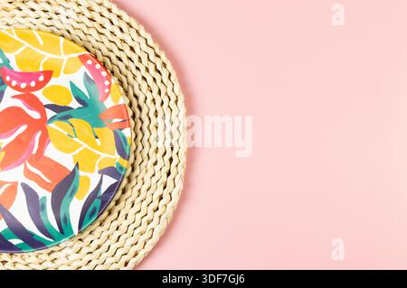 Top view of brown wicker table mat, colorful plate on pink background. Straw round tray, exotic plant plate, summer picnic. Flat lay, copy space. Stock Photo