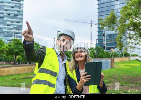 Architects and engineers review development plans on a tablet while pointing toward a construction site, collaborating on urban planning, smart city d Stock Photo