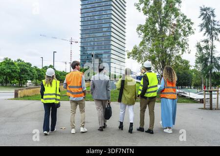 Diverse team of engineers, architects, and business executives standing with backs to camera, discussing the progress of a large construction project Stock Photo