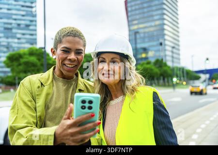 Diverse women architects and engineers smiling outdoors, looking at a smartphone screen, collaborating on an urban construction project, symbolizing t Stock Photo