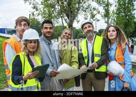 Multi ethnic team of engineers and architects in hard hats and safety vests reviewing blueprints on an urban construction site, collaborating on plann Stock Photo
