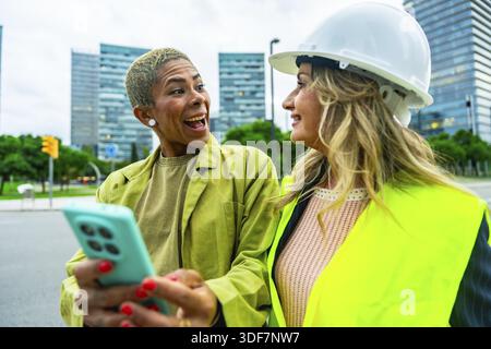 Two diverse colleagues. One an architect wearing a hardhat and safety vest. Looking at a smartphone held by her associate. Sharing a laugh while worki Stock Photo