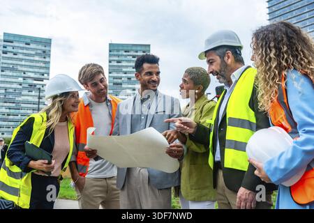 Diverse group of architects, engineers, and real estate professionals collaborating and discussing a blueprint at a construction site in a modern city Stock Photo