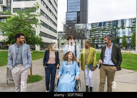 Diverse group of smiling business people, including a woman in a wheelchair, actively collaborating and communicating while walking through a contempo Stock Photo