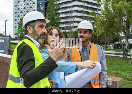 Group of diverse architects and engineers wearing hard hats and safety vests, discussing building plans and development while working outdoors in an u Stock Photo