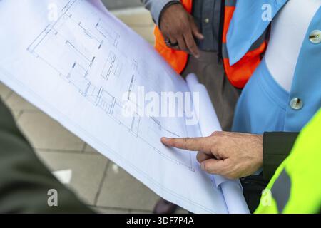 Engineers and architects in safety vests analyzing and discussing a detailed building blueprint, pointing at specific elements on the floor plan for c Stock Photo