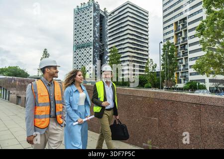 Diverse team of engineers and executives wearing safety vests and hard hats walking outdoors in a modern city street, discussing construction plans an Stock Photo