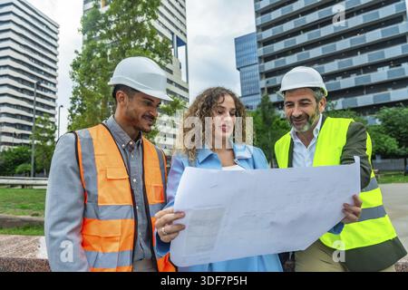 Construction team leader, architect, and engineer collaborating on a project, examining building plans and discussing ideas while standing in an urban Stock Photo