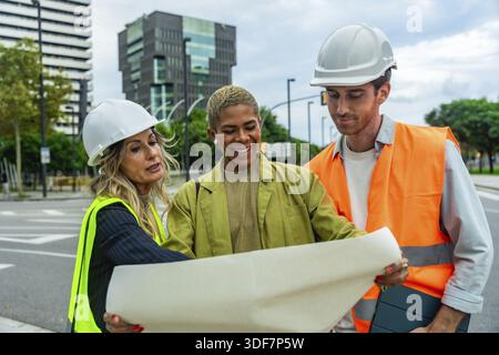 Diverse group of engineers and architects collaborating on an outdoor construction site, discussing project plans, safety, and urban development while Stock Photo