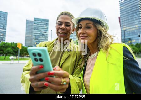 Diverse women architects and engineers smiling and collaborating outdoors in a modern city, using a smartphone for teamwork and communication on a con Stock Photo