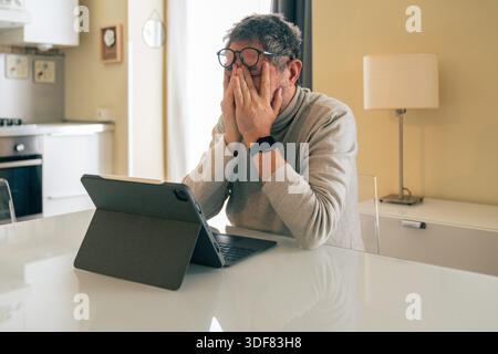 Senior man sitting at a table at home with a digital tablet, covering his face with his hands. Concept of mental fatigue, stress, emotional overwhelm, Stock Photo