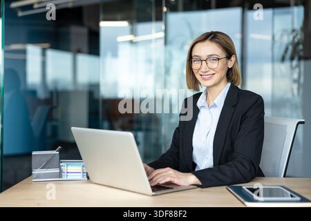 Smiling businesswoman wearing eyeglasses and a suit, sitting at a desk with a laptop, actively working and projecting confidence in a modern corporate Stock Photo