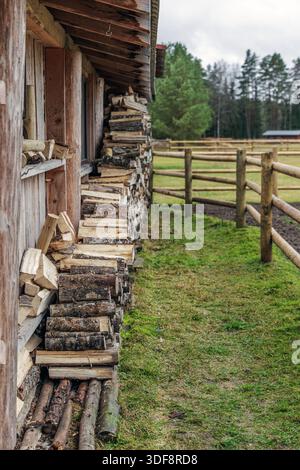 Neatly stacked logs of firewood under the eaves of a wooden farm building next to a green field and fence. Stock Photo