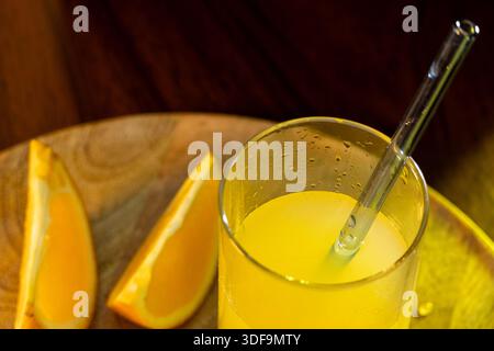 A glass of freshly squeezed orange juice with a glass straw and orange slices and halves lying next to it on a wooden tray. Stock Photo