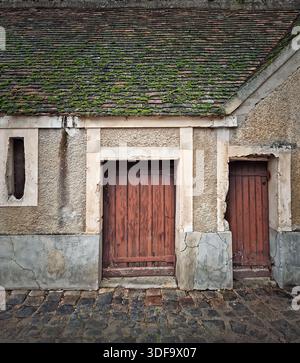 Rustic building with moss covered tiled roof and textured walls. Two old wooden doors with vertical planks next to a narrow window. Cobblestone street Stock Photo