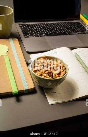 Vertical close-up view on office desk: bowl of walnuts, open weekly planner, notebook, pen, and laptop. Concept of healthy snacking, brain food for st Stock Photo