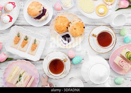 Easter or spring tea table scene against a white wood background. Selection of finger sandwiches, scones and sweet desserts. Overhead view. Stock Photo