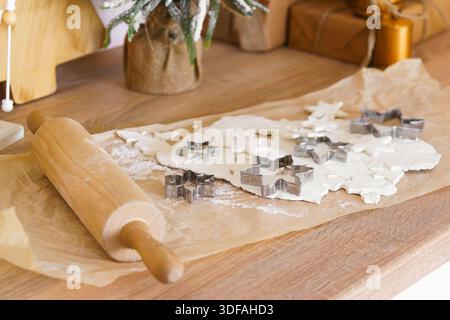 Festive Christmas baking process with rolled dough, metal cookie cutters for gingerbread and wooden rolling pin on parchment paper. Holiday kitchen sc Stock Photo