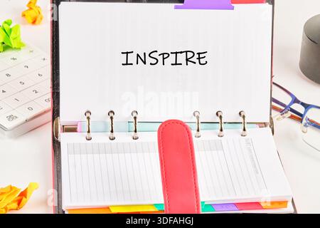 An open planner reads the word inspire, sitting amid crumpled paper, a calculator, glasses, and other desk items on a white surface, creating an atmos Stock Photo