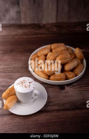 a cozy rustic breakfast scene: white porcelain cup of frothy cappuccino with chocolate sprinkles on a saucer, paired with a ceramic bowl piled high wi Stock Photo