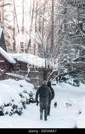 A couple with dogs walking along a snowy path toward a cozy cabin surrounded by winter forest during gentle snowfall Stock Photo
