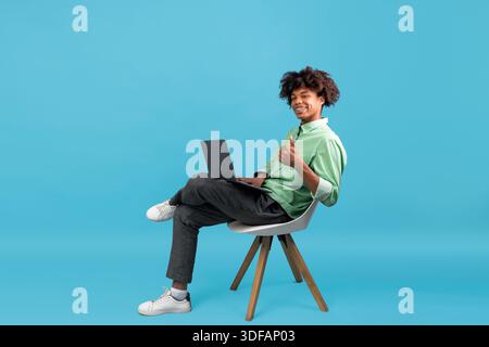 Happy black teen guy sitting with laptop in chair and showing thumb up, recommending online courses, blue background Stock Photo