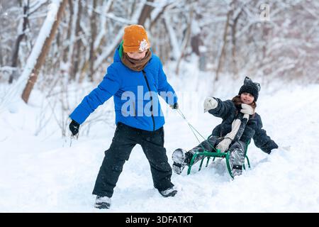 Boy pulling girl on sled during winter walk outdoors. kids enjoying winter activities in snowy forest. winter holidays, active childhood, screen-free Stock Photo
