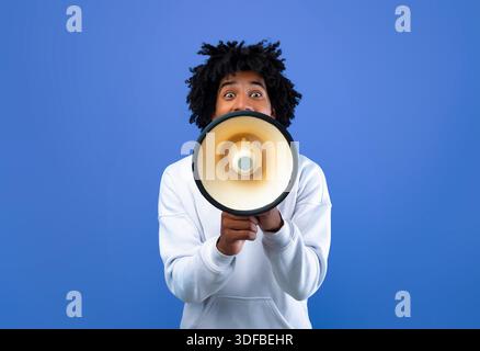 Emotional black teenager making announcement with megaphone on blue studio background Stock Photo