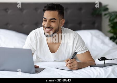 Young Arab Man Studies Online in Bed While Taking Notes and Smiling at the Laptop Screen Stock Photo