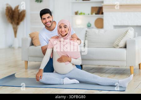 Pregnancy workout. Happy arab spouses exercising at home, woman stretching legs while man massaging wife's shoulders Stock Photo