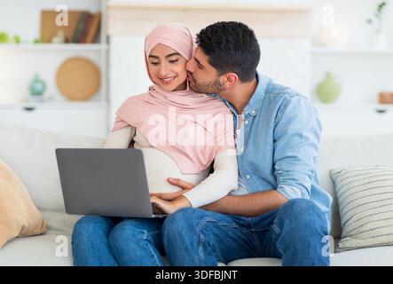 Muslim expecting woman and her loving husband using laptop, relaxing on sofa and searching furniture for baby room Stock Photo