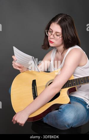 Musician is puzzled, holding musical notes in front as she figures out and breaks down new piece of music. Singer songwriter with guitar on lap. She i Stock Photo