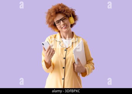 Redhead female freelancer in pajamas with gadgets on lilac background Stock Photo