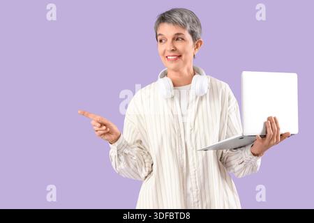 Mature woman with laptop pointing at something on lilac background Stock Photo