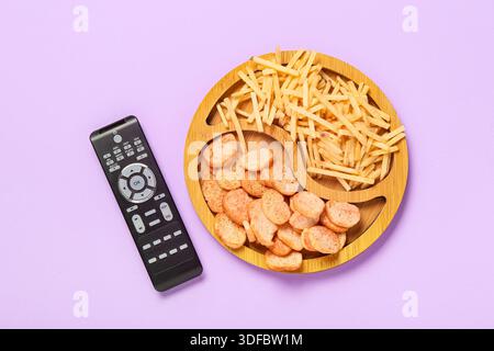 Wooden plate with crispy snacks and modern TV remote control on purple background Stock Photo