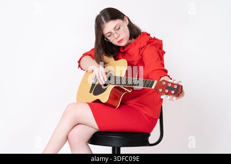 Woman guitarist tuning spanish acoustic guitar, sits on bar stool and looking at string with focus and concentration. Routine creative process of tuni Stock Photo