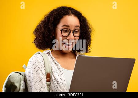 Young Woman Shows Excitement While Using Laptop With Yellow Background and Backpack Stock Photo