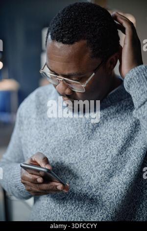 Middle Aged Black Man Using Smartphone While Scratching Head Indoors Stock Photo