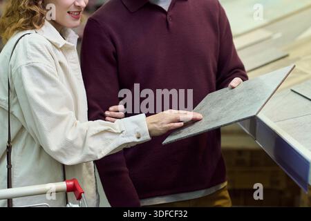Caucasian young adult woman and Caucasian young adult man examining ceramic tile samples together in store, woman touching surface while man standing beside her, both partially visible Stock Photo