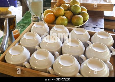 Refreshing morning setup featuring stack white ceramic coffee cups organized in wooden tray near fresh tangerine oranges on table waiting for breakfas Stock Photo