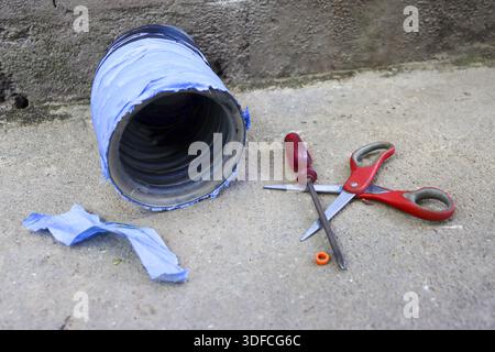 Old coffee can diy project wrapped in blue tape sitting on concrete floor next to red screwdriver and open pair of scissors during creative home repai Stock Photo