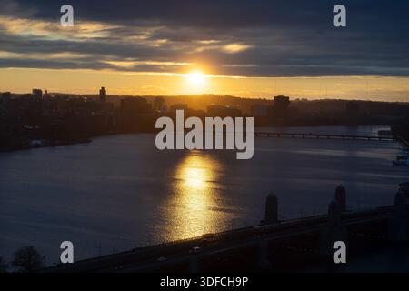 Sunset light reflects off the Charles River as the sun drops behind the skyline of Boston, with bridges silhouetted against the evening sky. Stock Photo