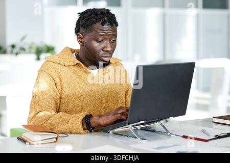 Young Adult Black Man Concentrating While Typing on Laptop at Office Desk Stock Photo