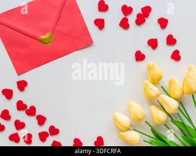Red envelope with heart sticker, yellow tulips and scattered felt hearts on white background. Valentines Day, love letter and romantic gift concept. Stock Photo