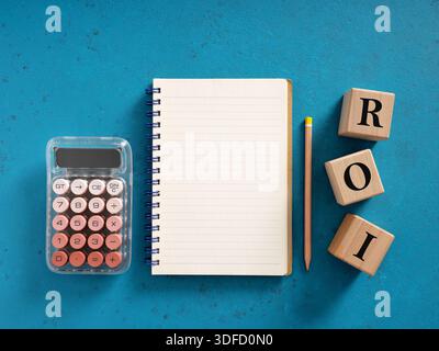 Return on investment business concept with wooden cubes showing financial ROI acronym, spiral notepad, pencil and calculator on blue office desk Stock Photo