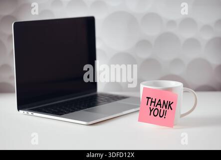 Thank you message is written on a sticky note sticked to a coffee cup on a white table near a laptop, gratitude concept Stock Photo