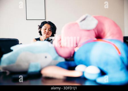 Asian woman with short curly hair sits at a table surrounded by colorful plush elephant toys. The scene is bright and playful, emphasizing a cozy atmo Stock Photo