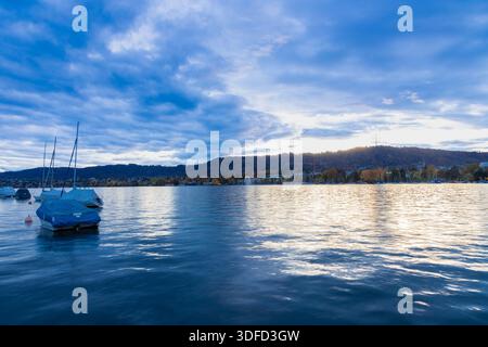 Sunset view during Fall over the famous lake Zurich with views on the autumn coloured pine trees on the Swiss Alps with some sunbeams and the boats an Stock Photo