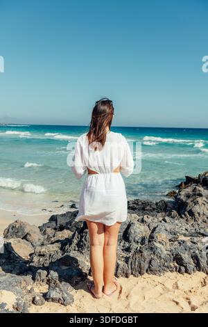Young woman in white dress looking at the ocean from a rocky beach Stock Photo