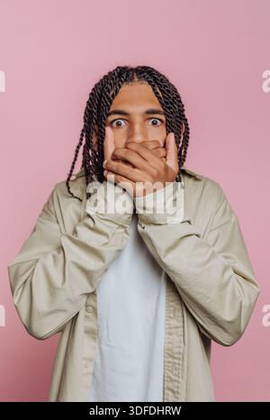 A young man stands with eyes wide open, showing surprise. His hands cover his mouth as he reacts to something unexpected. The background is a simple p Stock Photo